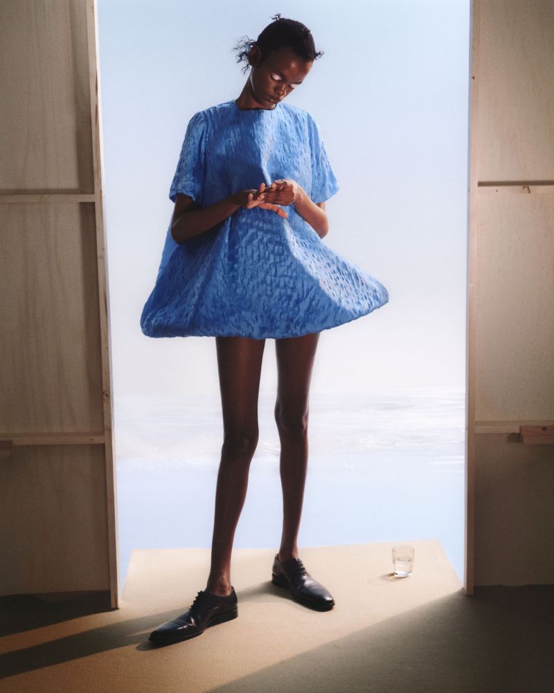 Fashion shot of a slender, dark-skinned woman in a voluminous blue dress and black shoes, standing next to a partially visible white wall.