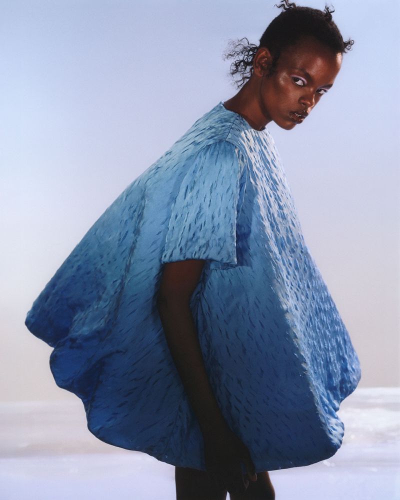 Fashion model wearing a draped, textural, light-blue dress posing in a studio with soft, diffused lighting against a graduated blue backdrop.