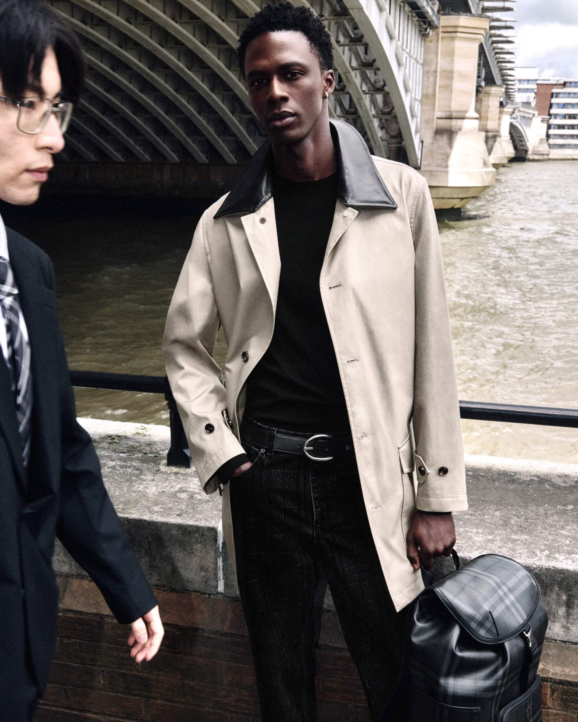 Black man wearing a beige coat with black collar standing by a bridge with water underneath holding a plaid backpack