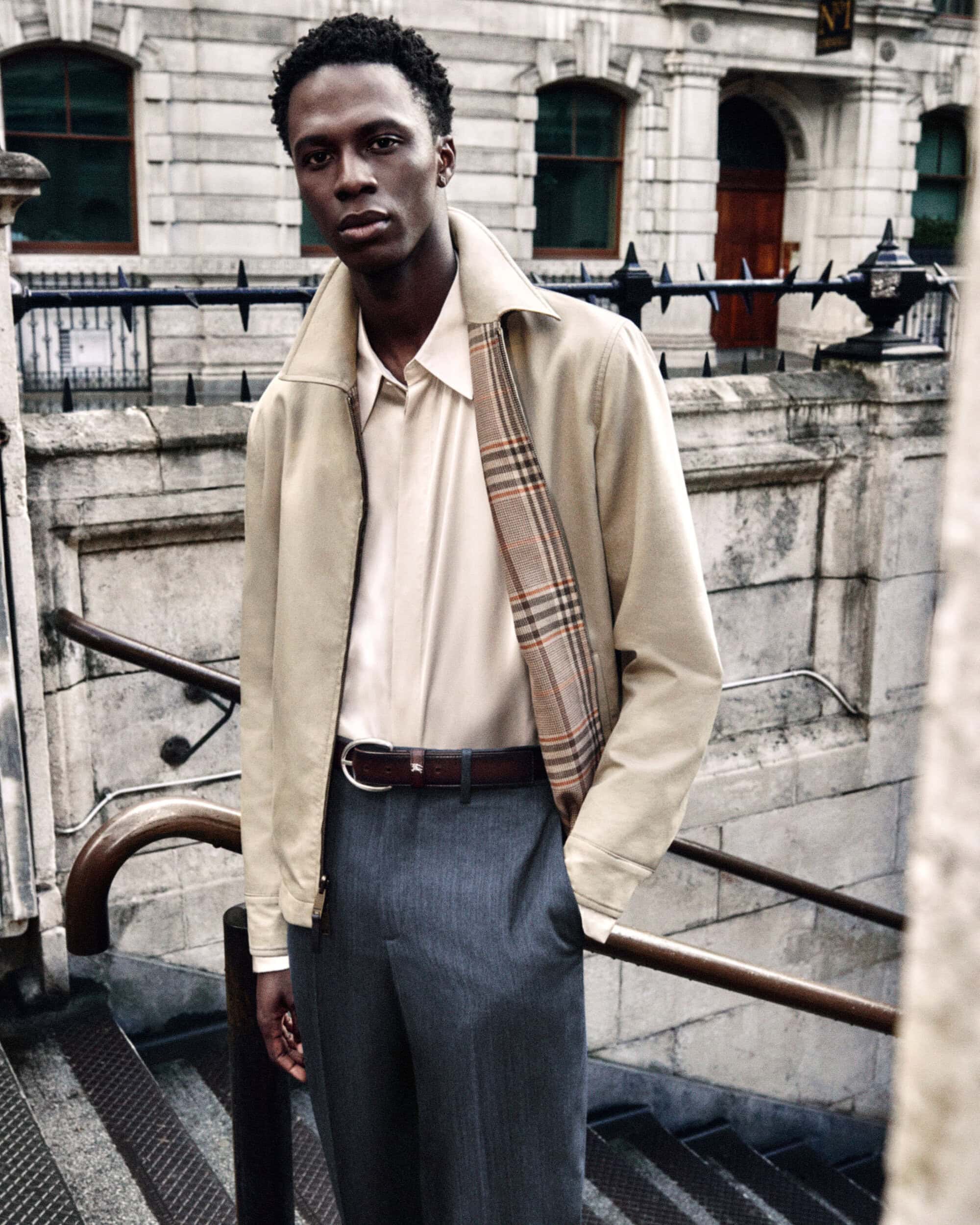 Black male model in a beige zip jacket with plaid lining, cream shirt, and grey trousers on outdoor stone steps.