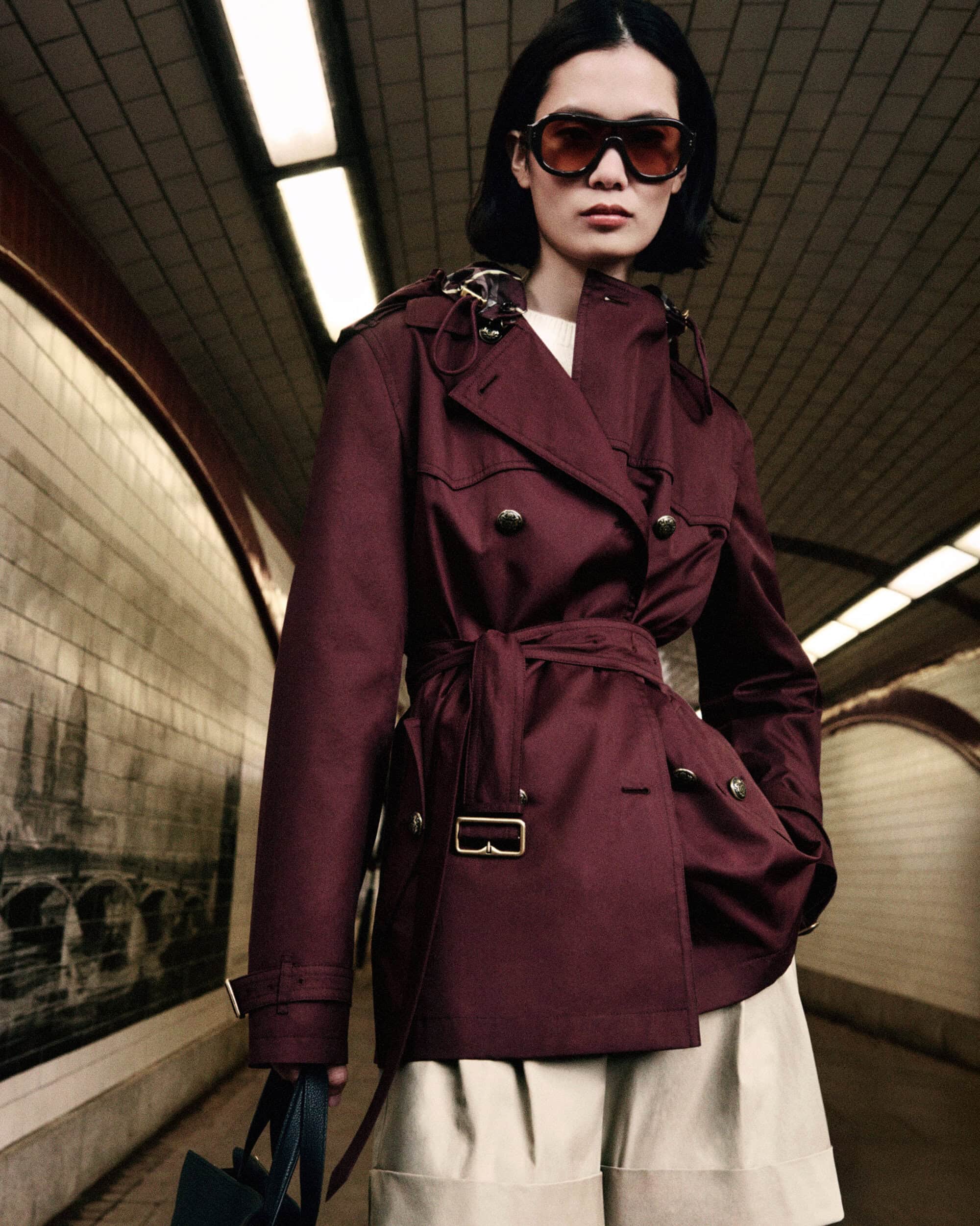 Model wearing a burgundy belted trench jacket and large sunglasses posing in a tiled subway corridor for Burberry.