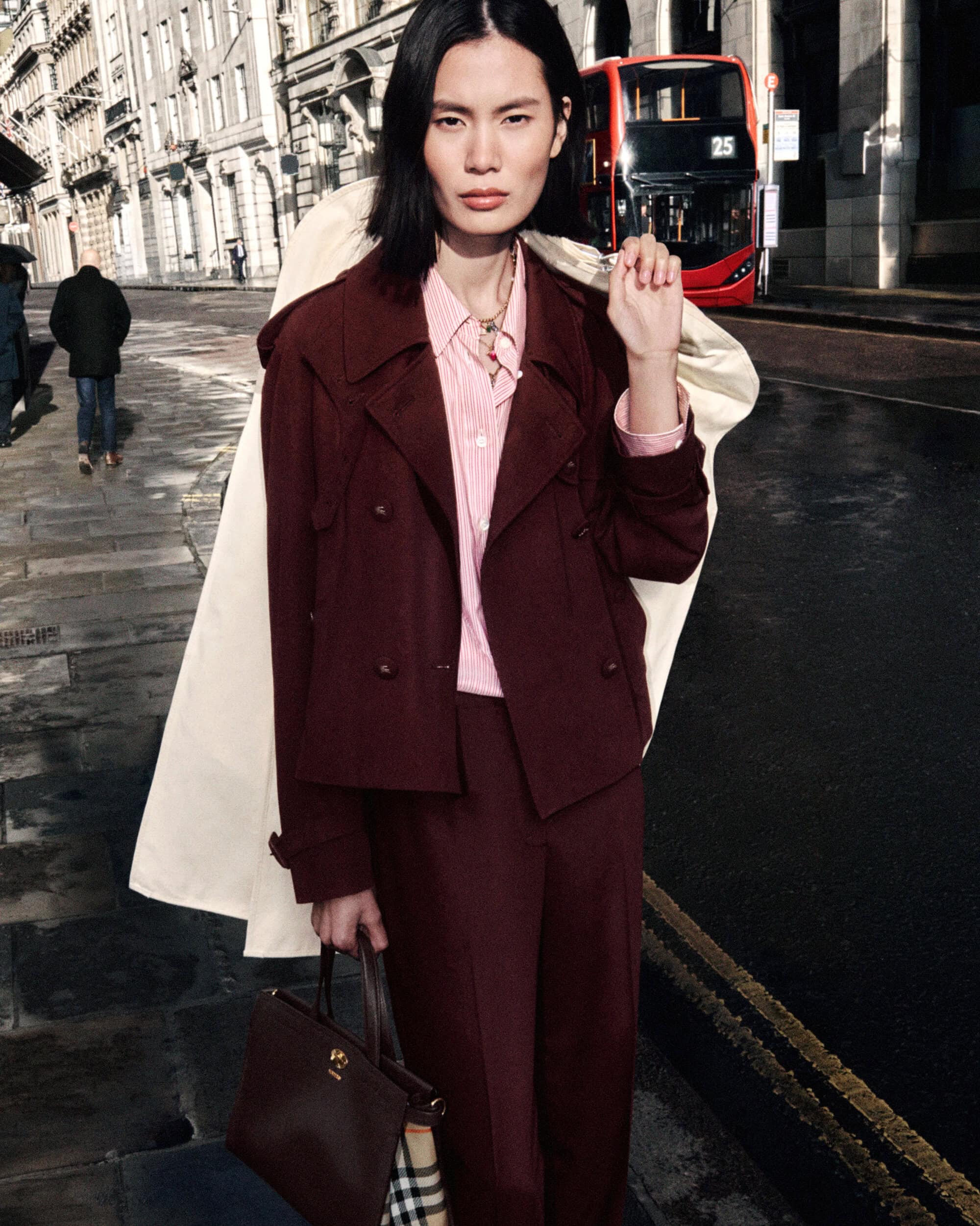 Model Haojie Qi wears a burgundy jacket and trousers with a pink shirt, holding a trench coat on a city street.