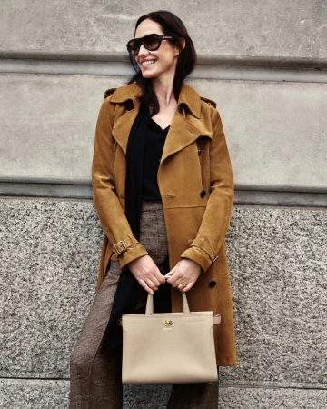 Rosemary Ferguson wearing a camel suede trench coat and sunglasses while holding a beige leather handbag against a stone wall.