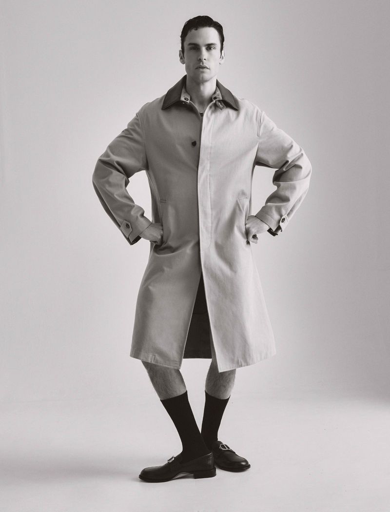 Man standing in beige trench coat with hands on hips, wearing black socks and loafers against a plain light gray studio background