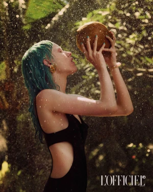 Model with teal hair in a black cut-out swimsuit holds a fresh coconut overhead in a sunlit shower of water droplets.
