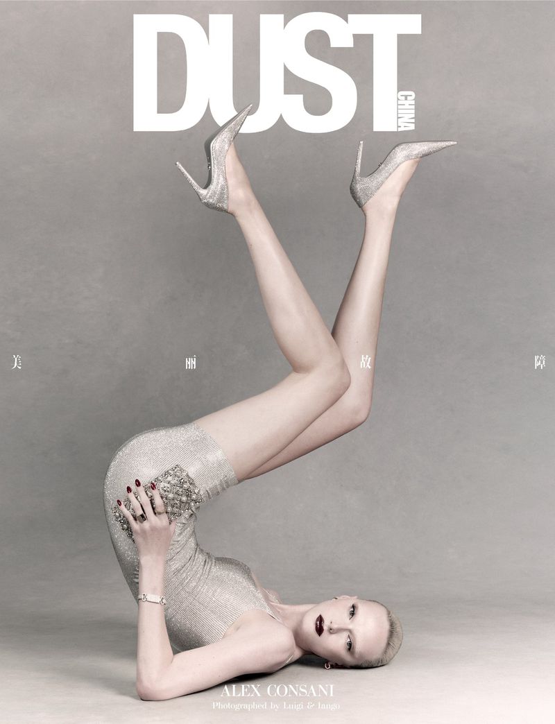 Model Alex Consani wearing a sparkly silver mini dress and heels, posing upside down on a studio floor with a silver clutch.