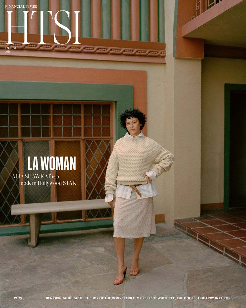 Actress Alia Shawkat wearing a textured cream sweater, white shirt, and midi skirt standing on a stone patio by a window.