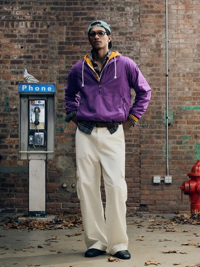 Man wearing a purple hooded windbreaker, layered plaid shirt, and cream trousers standing by an urban payphone with a perched pigeon.