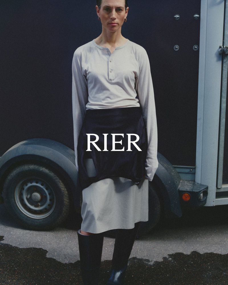 Model wearing long-sleeved light grey henley and skirt stands in front of a dark trailer in the RIER FW26 campaign.