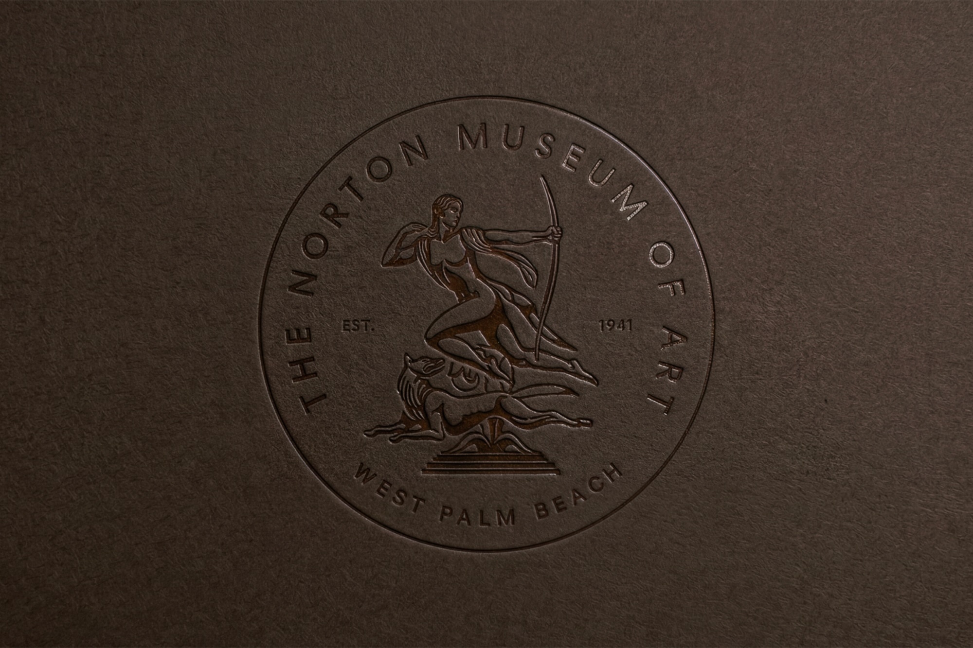 Embossed circular seal of The Norton Museum of Art featuring an archer and hound on dark brown textured paper.