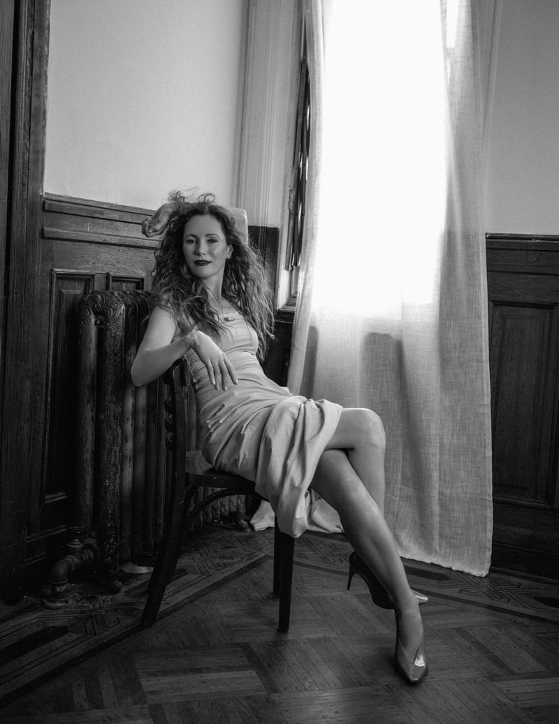 Leslie Mann seated on a wooden chair wearing a strapless midi dress and heels beside a bright curtained window in black and white.