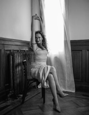 Leslie Mann seated on a wooden chair with arms raised overhead in a black and white room with wood paneling.