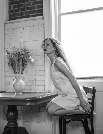Leslie Mann kneeling on a wooden chair in a white sleeveless dress beside a table with a flower vase in black and white.