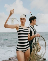 Model in a black and white striped swimsuit holds a water-filled bag with a perfume bottle on a rocky coast alongside a diver.