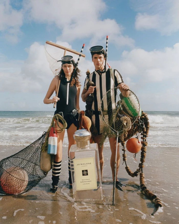 Two models in vintage black and white swimwear holding fishing nets behind an oversized Jo Malone perfume bottle on a beach.