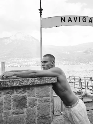 Monochrome portrait of model Josh McGregor shirtless, leaning his arms on a stone ledge near a lakeside pier.