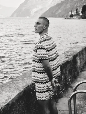 Model Josh McGregor in a patterned knit shirt and shorts standing beside a lake in black and white for Man About Town.