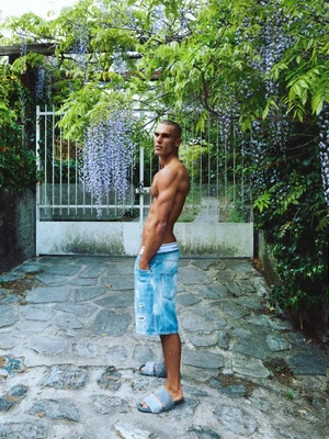 Josh McGregor posing shirtless in denim shorts and textured slides in front of a garden gate draped in purple wisteria.