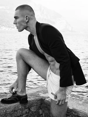 Model Josh McGregor in profile wearing an open black blazer and white shorts with studded brogues next to a lake.