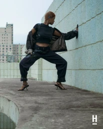 Kelela posing in a wide stance wearing an all-black streetwear outfit and kitten heels against a textured wall on an urban rooftop.
