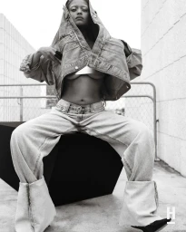 Kelela seated wearing an oversized denim jacket pulled over her head and wide-leg denim jeans in a black and white editorial portrait.