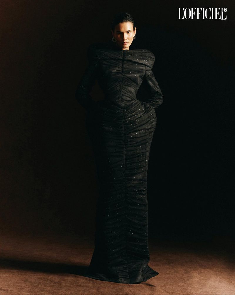 Woman in long black evening gown posing in a dimly lit studio for L'Officiel fashion editorial, elegant and sophisticated.