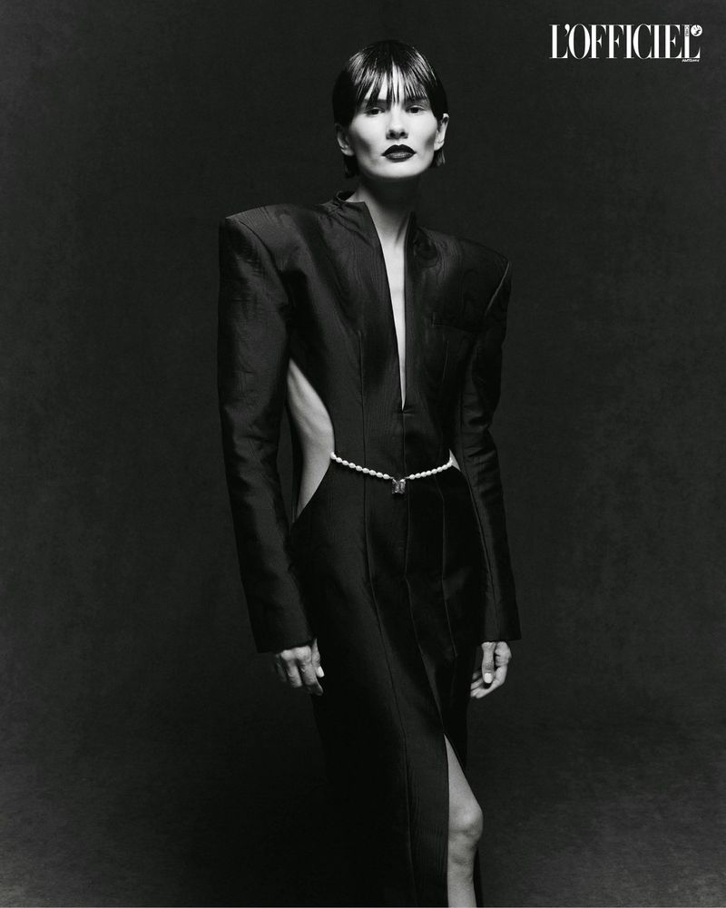 Elegant editorial fashion shot of a slender adult female with dark hair wearing a black dress and pearl belt against a dark backdrop.