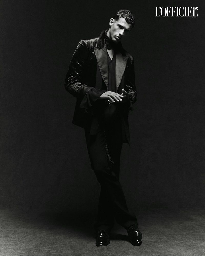 Black and white portrait of a man in a velvet suit and black shirt posing in a studio with dark background; fashion editorial photography.