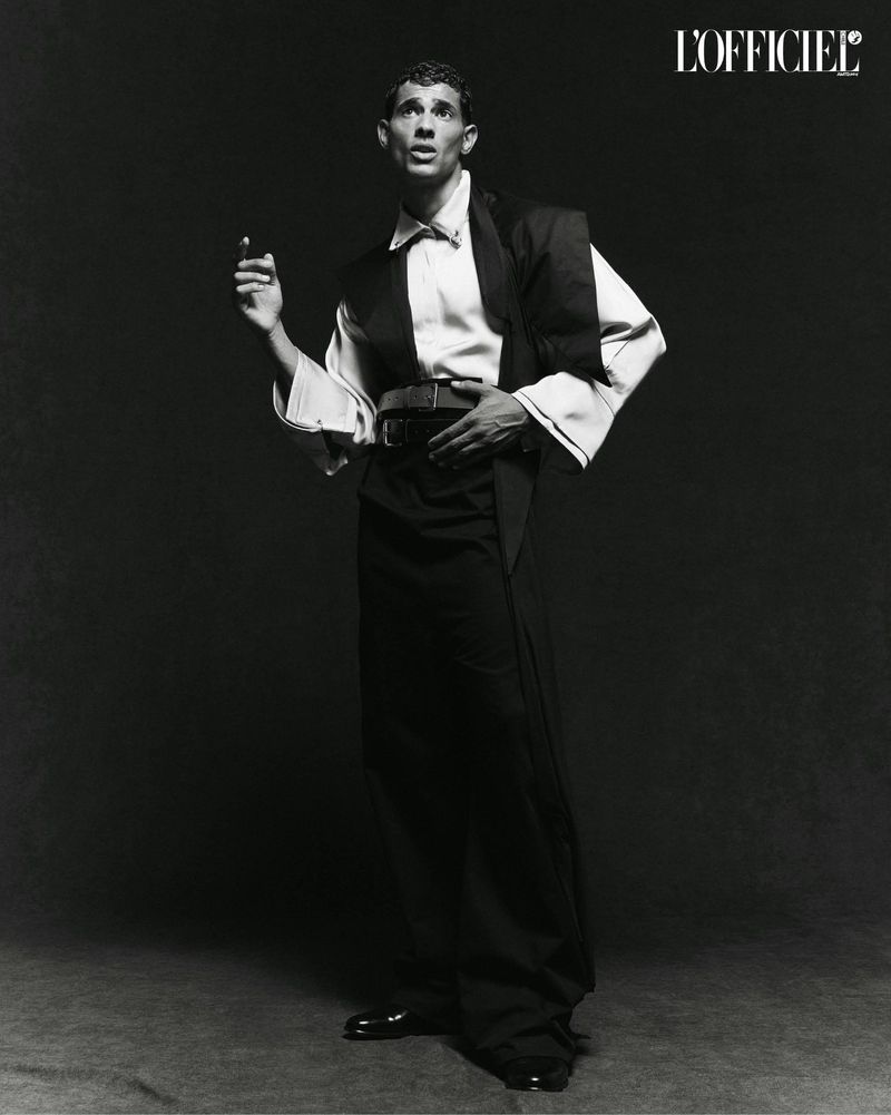 A man poses in a black and white studio portrait, wearing a wide-collared shirt, vest, and slacks with patent leather shoes and a belt.