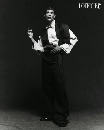 A man poses in a black and white studio portrait, wearing a wide-collared shirt, vest, and slacks with patent leather shoes and a belt.