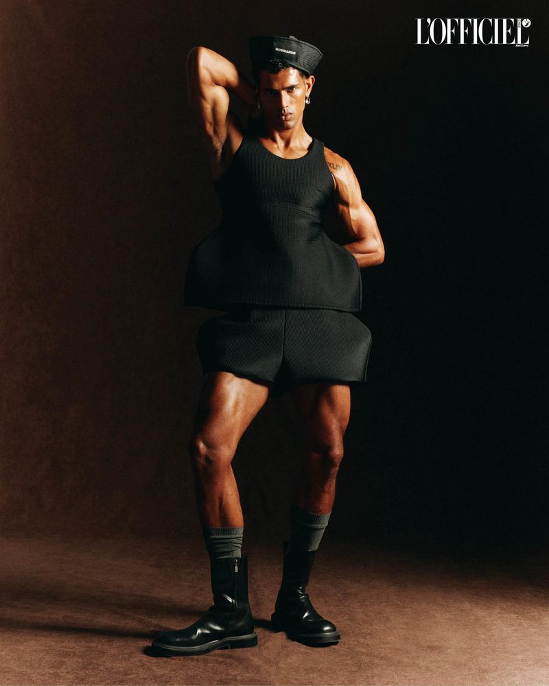 Muscular man posing in black outfit, hat, and boots in a studio setting with gradient brown and black background, fashion editorial.