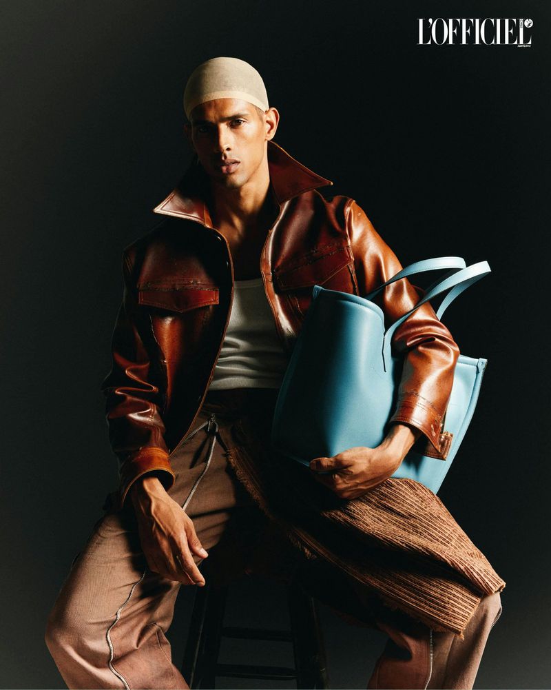 A model in a brown leather jacket sits indoors against a dark background, holding a light blue tote bag, in a fashion editorial shot.