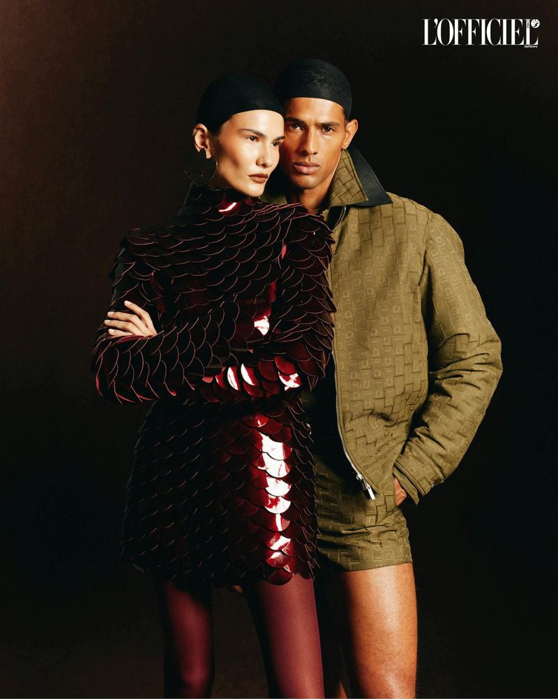 Editorial shot of a biracial couple modeling designer outfits in a dark studio setting for a fashion magazine cover, looking elegant and stylish.