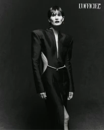 Model in a black structured dress with shoulder pads and a delicate waist chain against a dark textured backdrop