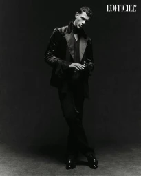 Black and white portrait of a male model wearing a dark velvet blazer and trousers against a textured dark background.
