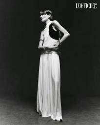 Model in a long white draped dress with black paneling standing against a dark studio backdrop for L'Officiel Chile.