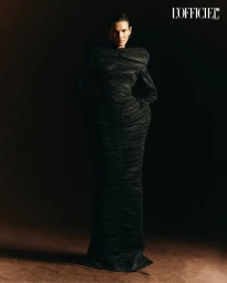 Model standing in a structured black gown against a dark textured background for L'Officiel Chile editorial photography.