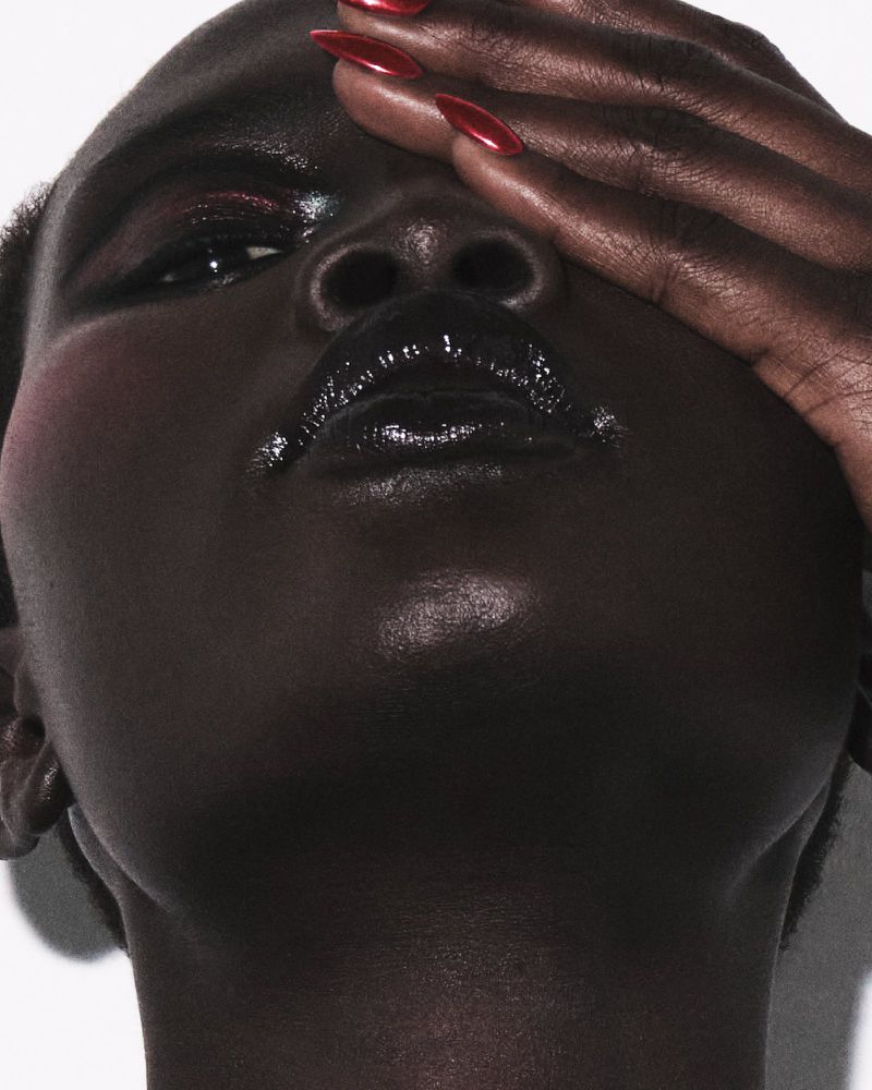 Close-up portrait of a model with glossy black lipstick and hand covering upper face showing glossy red nails