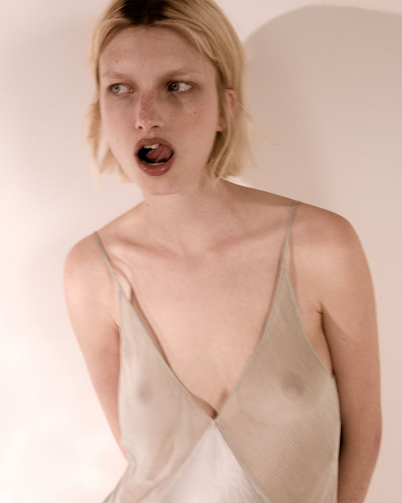 Model Kristin Drab in a sheer pale slip dress posing against an off-white wall with soft shadow.