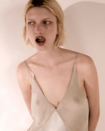 Model Kristin Drab in a sheer pale slip dress posing against an off-white wall with soft shadow.