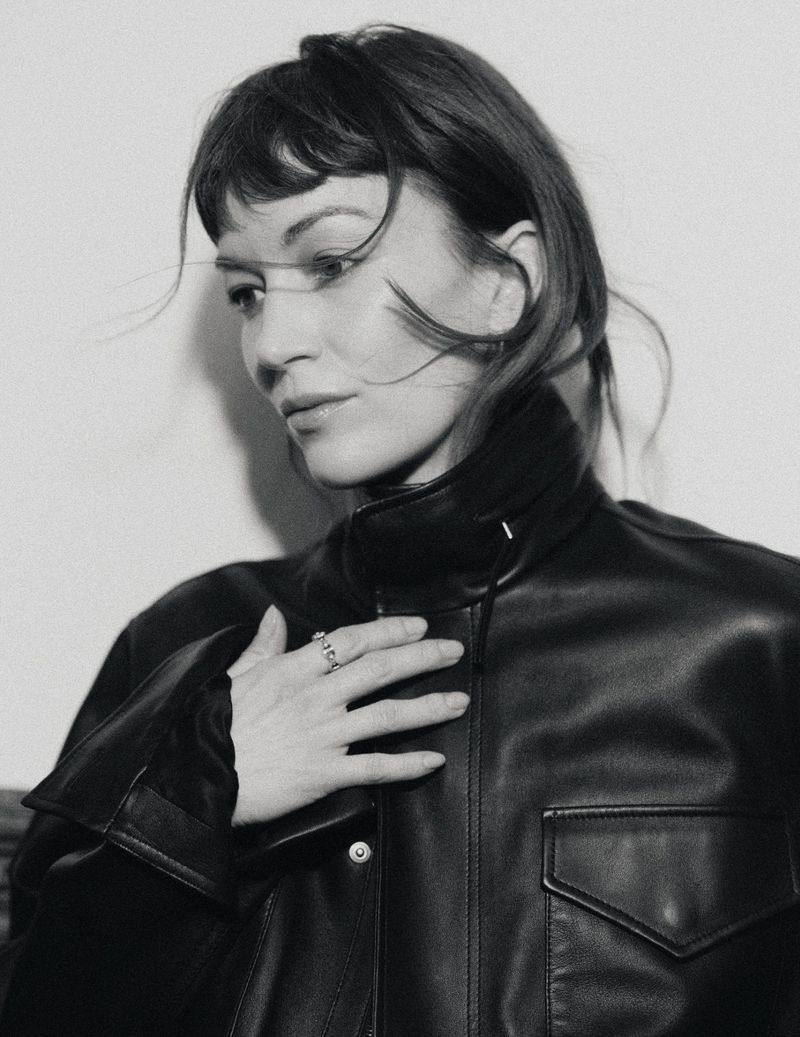 Portrait of actor Britt Lower wearing a black leather jacket, hands resting on chest, against a plain white background.