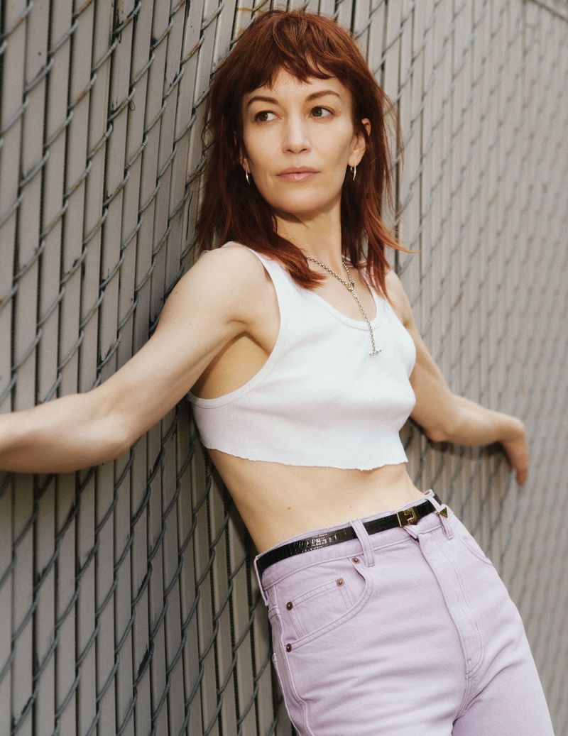 Britt Lower leaning against a chain-link fence wearing a white cropped tank top and lavender high-waisted jeans.