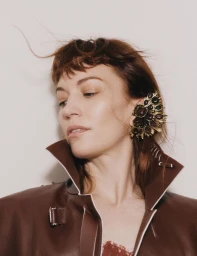Britt Lower wearing an ornate jeweled ear cuff and high-collared brown leather jacket against a seamless off-white background.