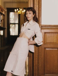 Britt Lower wearing a pale matching shirt and skirt leaning against a carved wooden doorframe in an interior space for Flaunt.