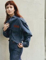 Britt Lower wearing a dark blue denim jacket and matching skirt against a neutral wall in an editorial portrait.