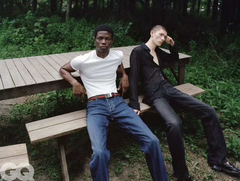 Two men wearing casual summer clothing seated on a wooden picnic table bench surrounded by lush green foliage.
