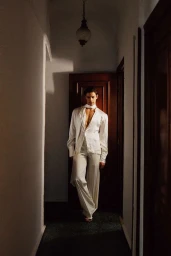 Model Francisco Rosso posing in an open white silk shirt and wide-leg trousers inside a dimly lit residential hallway.