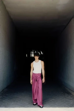 Model Francisco Rosso stands wearing a white ribbed tank top and draped mauve wide-leg trousers in a dark concrete passageway.