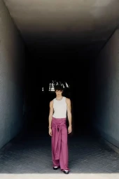 Model Francisco Rosso stands wearing a white ribbed tank top and draped mauve wide-leg trousers in a dark concrete passageway.