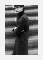 Model Francisco Rosso standing in profile wearing a tailored black coat against a corrugated metal wall in black and white.